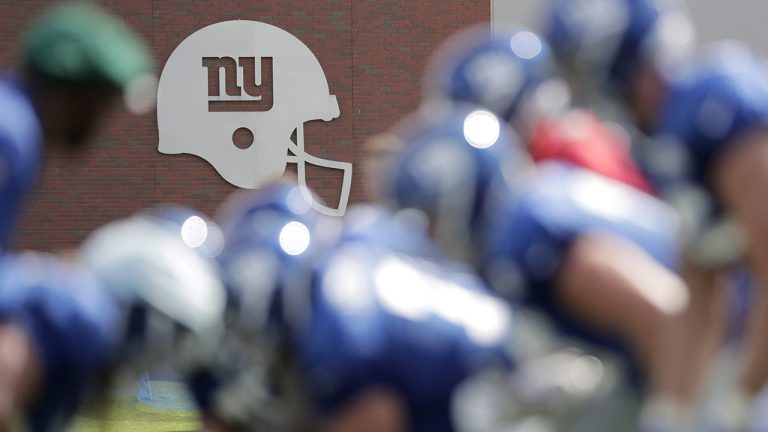 A logo on the side of a building is seen as New York Giants offensive players line up during NFL football training camp, Wednesday. (Julio Cortez/AP)