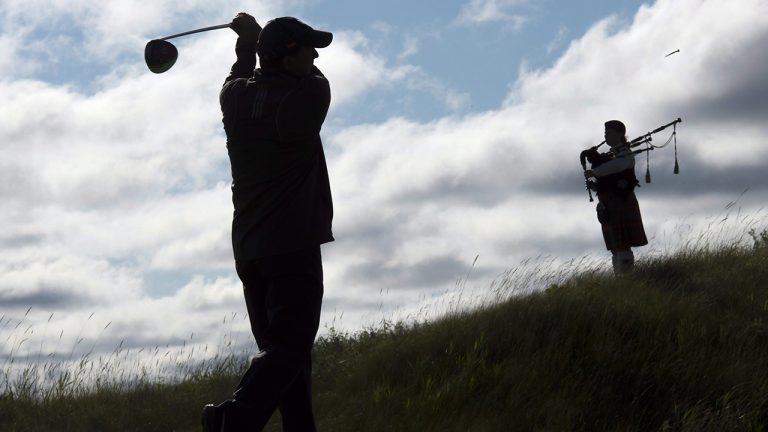 Golf course architect Doug Carrick tees off as bagpiper Kristen MacKay stands by the first tee during the Osprey Valley Heathlands 25th anniversary celebration in Caledon, Ont. (Frank Gunn/CP)