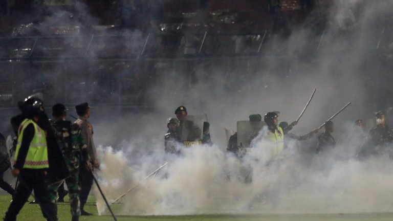 Police officers and soldiers stand amid tear gas smoke during a soccer match at Kanjuruhan Stadium in Malang, East Java, Indonesia, Saturday, Oct. 1, 2022. Clashes between supporters of two Indonesian soccer teams in East Java province killed over 100 fans and a number of police officers, mostly trampled to death, police said Sunday. (Yudha Prabowo/AP)