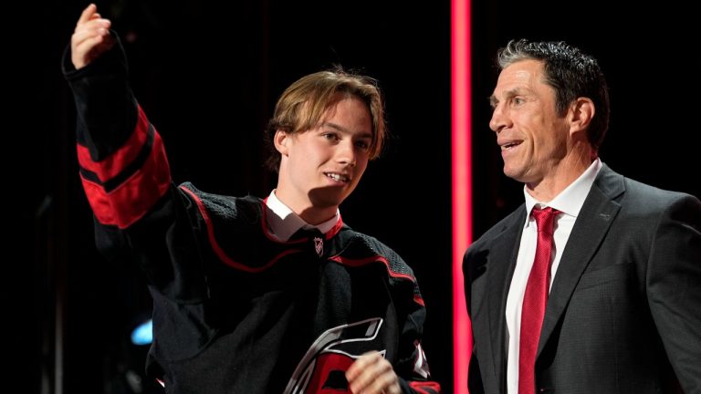 Bradly Nadeau puts on a Carolina Hurricanes jersey next to coach Rod Brind'Amour after being picked by the team during the first round of the NHL hockey draft Wednesday, June 28, 2023, in Nashville, Tenn. (AP Photo/George Walker IV)