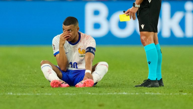 Kylian Mbappe of France sits on the pitch, hand over his bloody nose, as the referee holds the yellow card, during a Group D match between Austria and France at the Euro 2024 soccer tournament. (Andreea Alexandru/AP) 