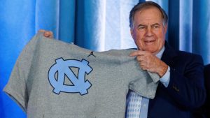 New North Carolina football coach Bill Belichick holds up a UNC branded sleeveless hoodie presented to him during an NCAA college football press conference announcing his hiring, Thursday, Dec. 12, 2024, in Chapel Hill, N.C. (AP Photo/Ben McKeown)